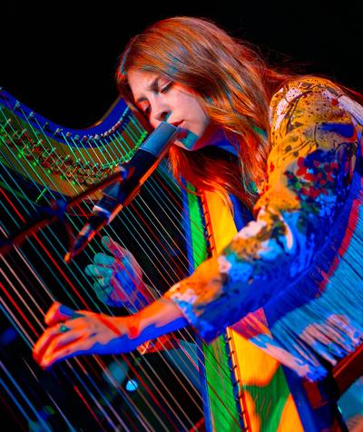 Mikaela Davis sings into a microphone while playing harp under vivid red, blue, and green stage lights.