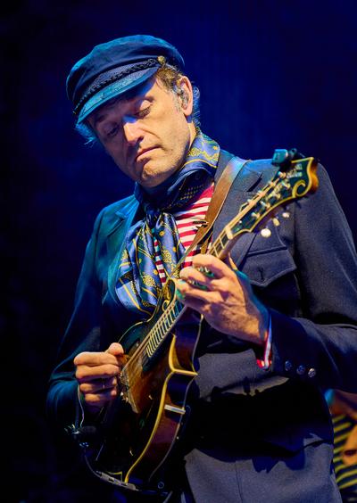 A member of Punch Brothers plays mandolin under blue stage lights, wearing a peaked cap and patterned scarf.