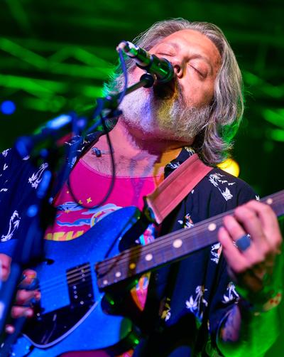 A member of The Dear Hunter sings into a microphone while playing a blue electric guitar under green stage lights.