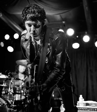 A Torturetwinn guitarist leans over their instrument in a black leather jacket in a black-and-white concert photograph.