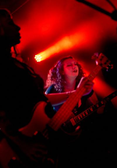 Members of Faetooth perform beneath deep red stage lights, framed by a silhouetted bassist in the foreground.